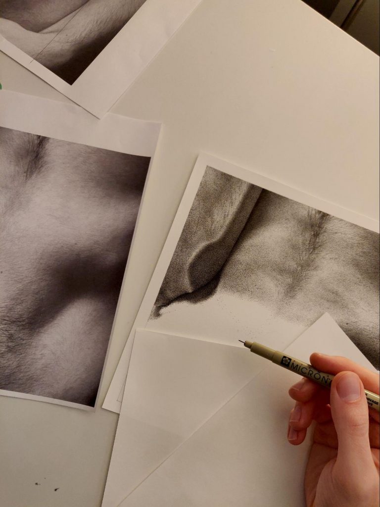 Justin Mezzapelli's hand holds a pen close to paper, about to illustrate. Three drawings on paper are scattered on a white surface while two blank pieces of paper lie below the hand, holding a pen.
